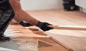 Man con guantes negros coloca una tabla de madera para reparar tarima flotante desgastada. Adhesivo aplicado en el suelo.