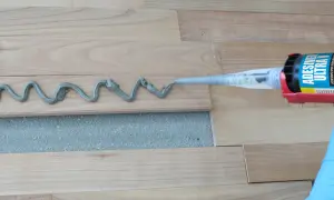 Aplicando adhesivo para rellenar juntas de parquet. Un guante azul sostiene una pistola de calafateo que deposita una línea ondulada de material gris sobre las tablas de madera.