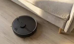 Robot aspirador negro en suelo de madera, realizando la limpieza bajo un sofá beige.