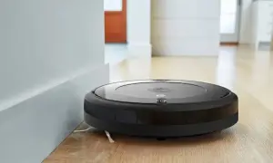 Robot aspirador Roomba negro junto a una pared blanca, listo para resetear y limpiar el suelo de madera.