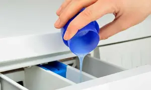 Mano vierte suavizante líquido de un tapón azul en el cajetín de la lavadora, preparándose para el lavado.