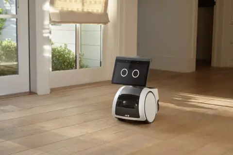 El robot Alexa Astro, con pantalla y ruedas, se encuentra en un suelo de madera, cerca de una puerta acristalada.