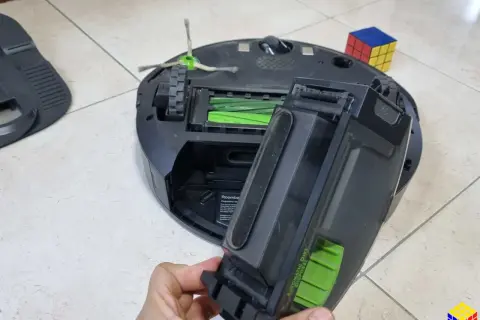 Mano retirando el depósito de suciedad de un Roomba, quizás para solucionar un error 11. Un cubo de Rubik descansa cerca.