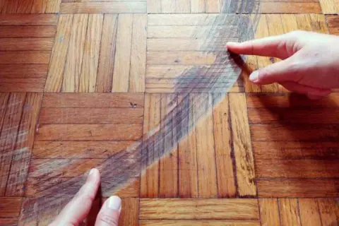 Manos señalan un arañazo profundo en un suelo de parquet de madera. Descubre cómo quitar arañazos del parquet y restaurar su brillo.