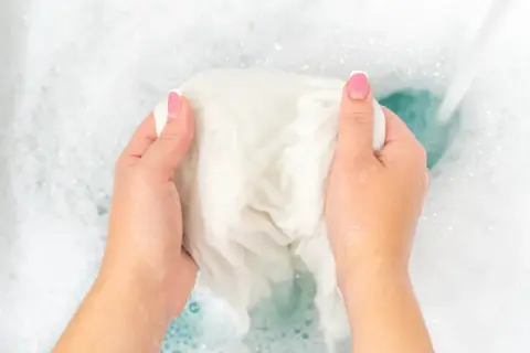 Manos lavando ropa blanca en agua con burbujas. Un chorro de agua cae sobre la tela, ayudando a como blanquear ropa blanca.