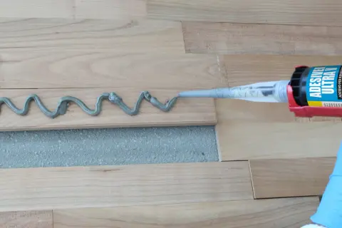 Aplicando adhesivo para rellenar juntas de parquet. Un guante azul sostiene una pistola de calafateo que deposita una línea ondulada de material gris sobre las tablas de madera.