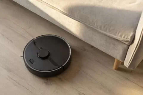 Robot aspirador negro en suelo de madera, realizando la limpieza bajo un sofá beige.