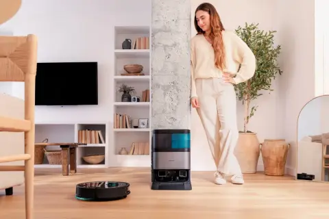 Una mujer sonríe junto a su nueva conga, un robot aspirador que limpia el suelo mientras ella descansa.