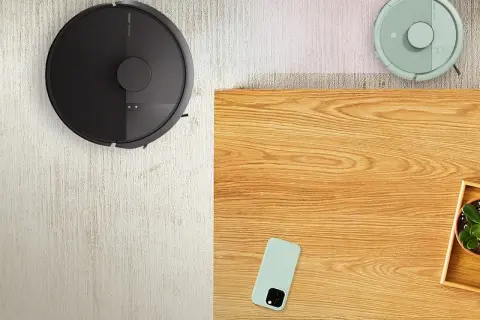 Dos robots aspiradores, uno negro y otro verde, limpian la alfombra. El negro parece tener una irobot bolsa, listo para recoger el polvo.