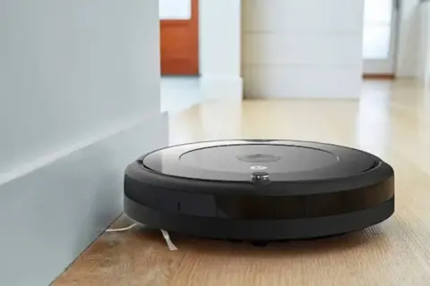 Robot aspirador Roomba negro junto a una pared blanca, listo para resetear y limpiar el suelo de madera.