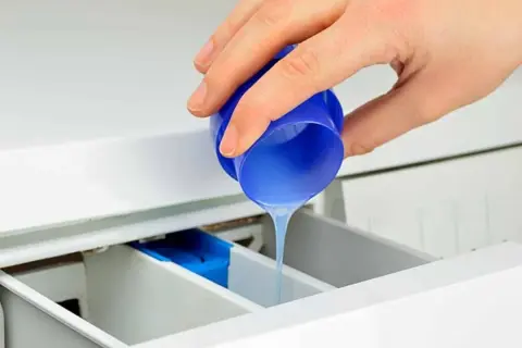 Mano vierte suavizante líquido de un tapón azul en el cajetín de la lavadora, preparándose para el lavado.