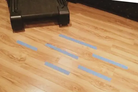 Cinta azul marca zonas de posible humedad debajo de la tarima. Un mueble oscuro está en la parte superior.