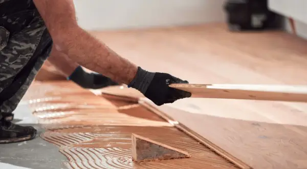 Man con guantes negros coloca una tabla de madera para reparar tarima flotante desgastada. Adhesivo aplicado en el suelo.