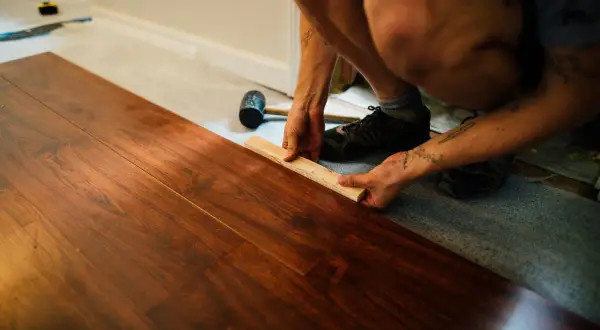 Instalando tarima flotante sobre parquet. Se muestra cómo colocar una pieza de madera con un bloque y mazo. El precio de poner tarima encima de parquet es una inversión que renueva el hogar.