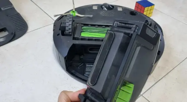 Mano retirando el depósito de suciedad de un Roomba, quizás para solucionar un error 11. Un cubo de Rubik descansa cerca.