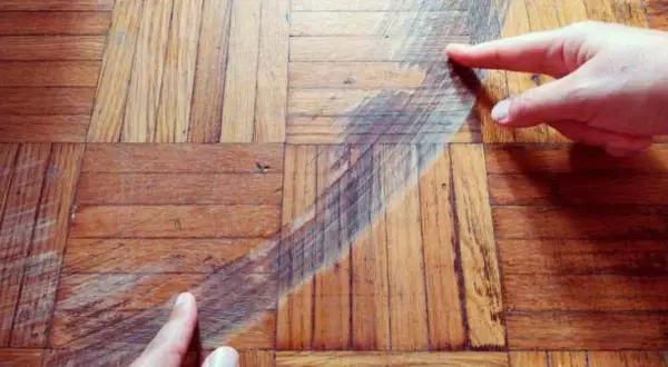 Manos señalan un arañazo profundo en un suelo de parquet de madera. Descubre cómo quitar arañazos del parquet y restaurar su brillo.