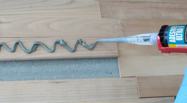 Aplicando adhesivo para rellenar juntas de parquet. Un guante azul sostiene una pistola de calafateo que deposita una línea ondulada de material gris sobre las tablas de madera.