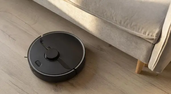 Robot aspirador negro en suelo de madera, realizando la limpieza bajo un sofá beige.