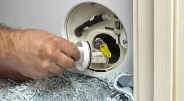 Mano retirando filtro sucio de lavadora que pierde agua por debajo. Se ve pelusa y un objeto amarillo.