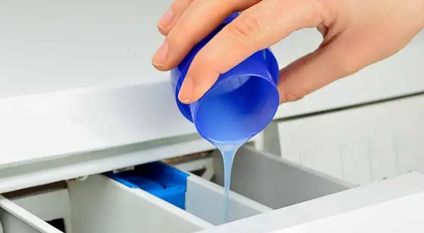 Mano vierte suavizante líquido de un tapón azul en el cajetín de la lavadora, preparándose para el lavado.