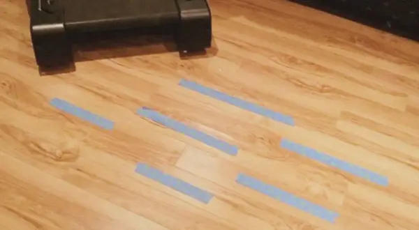 Cinta azul marca zonas de posible humedad debajo de la tarima. Un mueble oscuro está en la parte superior.