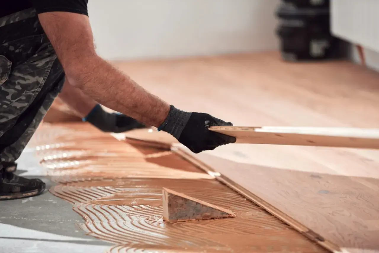 Man con guantes negros coloca una tabla de madera para reparar tarima flotante desgastada. Adhesivo aplicado en el suelo.