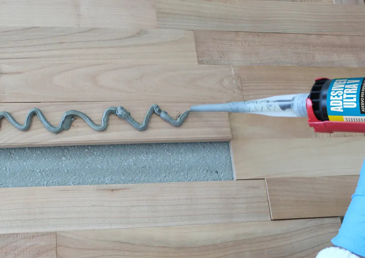 Aplicando adhesivo para rellenar juntas de parquet. Un guante azul sostiene una pistola de calafateo que deposita una línea ondulada de material gris sobre las tablas de madera.