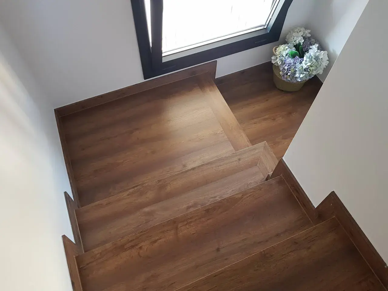 Escaleras con suelo laminado de madera, con un cesto de flores junto a una ventana.