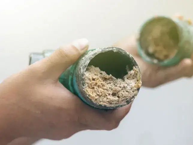 Manos sostienen tuberías obstruidas con depósitos calcáreos, un problema común que un desatascador casero podría solucionar.