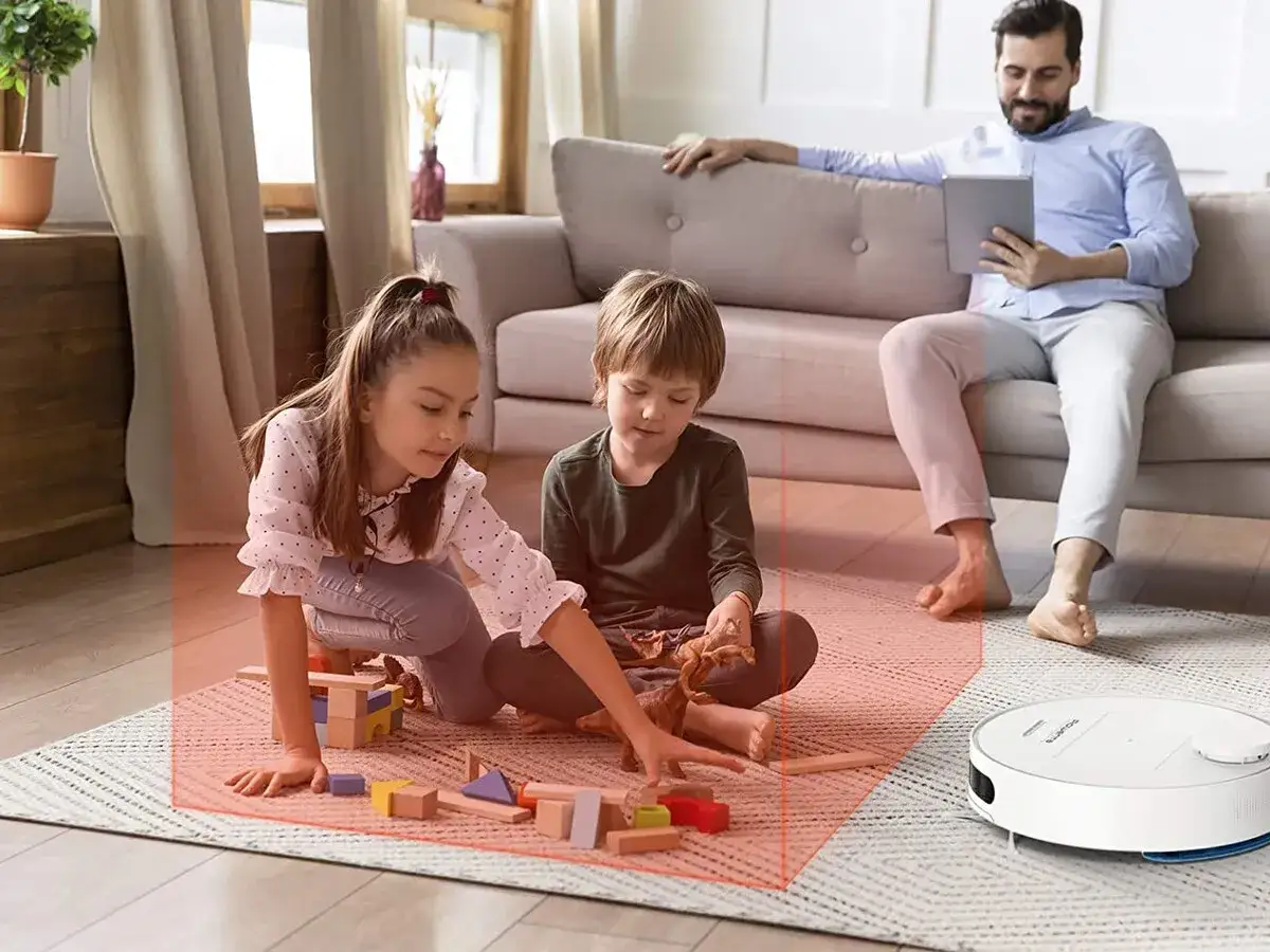 Un padre mira su tablet mientras sus hijos juegan en el suelo. El robot aspirador Rowenta Smart Force Essential se acerca, manteniendo la zona de juego despejada.