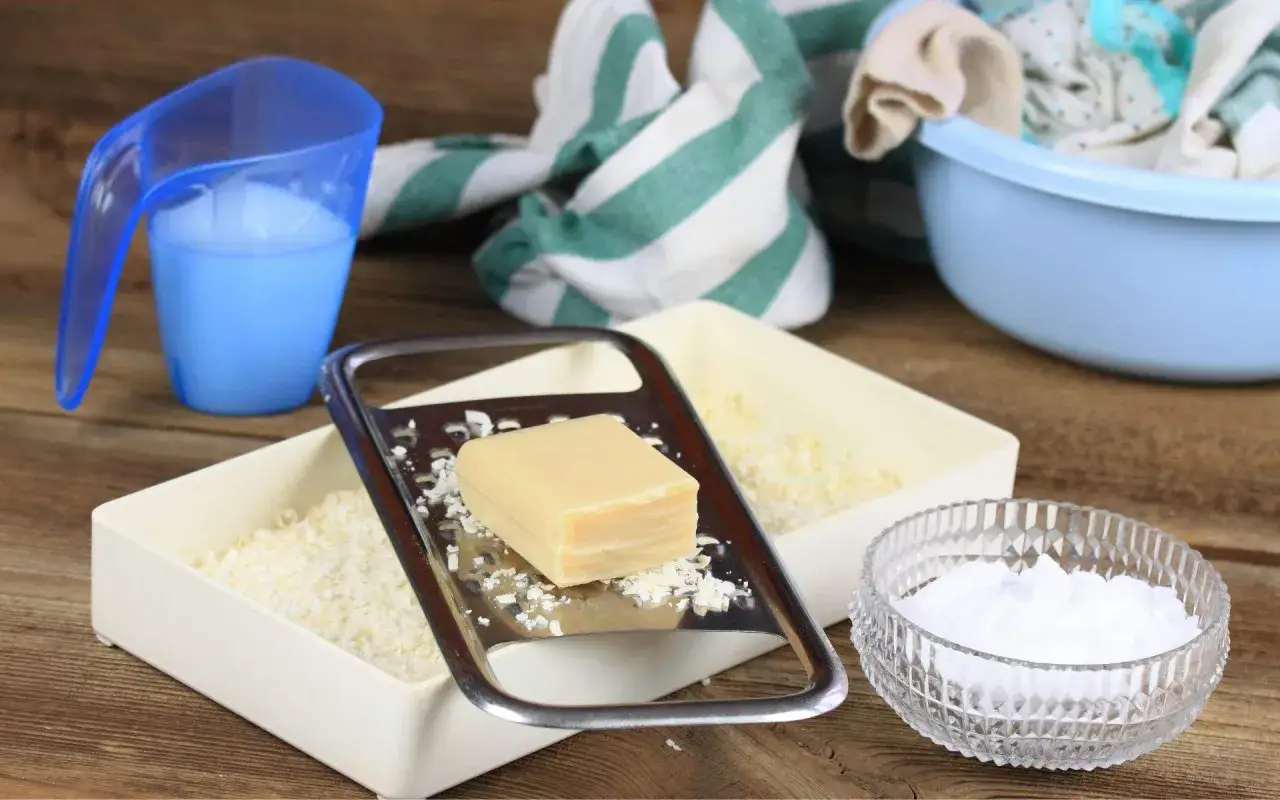 Preparando jab&oacute;n casero para lavadora: rallador con barra de jab&oacute;n, recipiente con virutas, taza medidora con l&iacute;quido y taz&oacute;n con ropa sucia.
