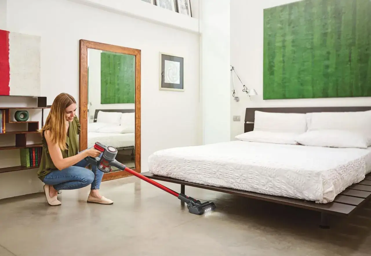 Mujer sonríe mientras usa la aspiradora Polti para limpiar el suelo de un dormitorio moderno.
