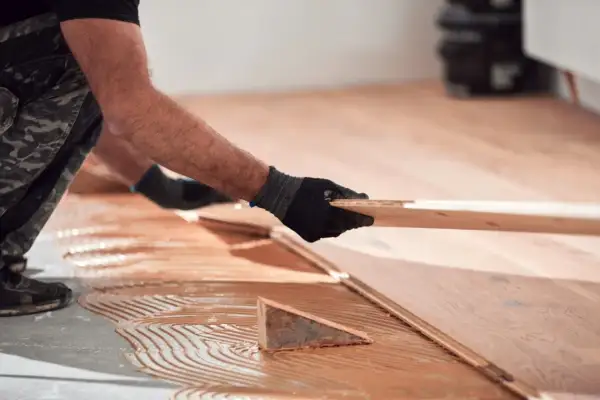 Man con guantes negros coloca una tabla de madera para reparar tarima flotante desgastada. Adhesivo aplicado en el suelo.
