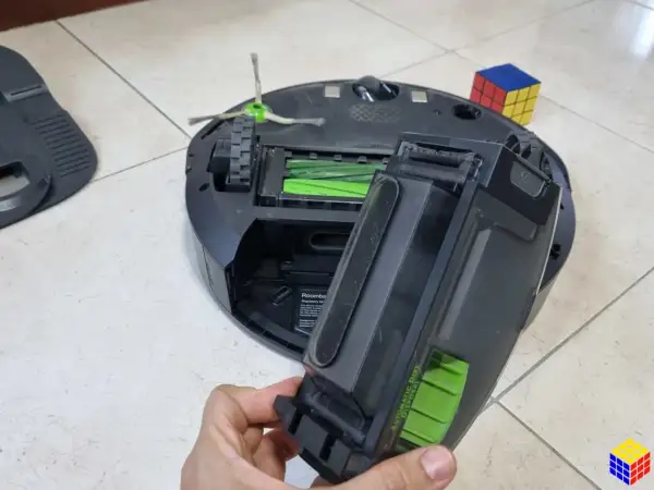 Mano retirando el depósito de suciedad de un Roomba, quizás para solucionar un error 11. Un cubo de Rubik descansa cerca.