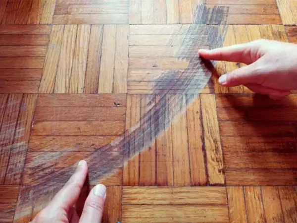Manos señalan un arañazo profundo en un suelo de parquet de madera. Descubre cómo quitar arañazos del parquet y restaurar su brillo.
