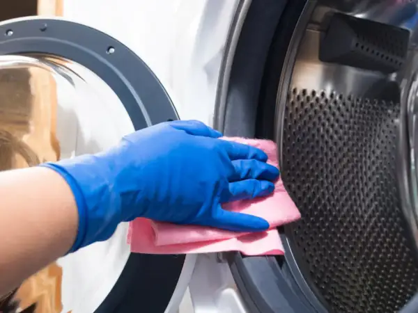 Mano con guante azul limpia el interior de una lavadora, eliminando una mancha de ropa.